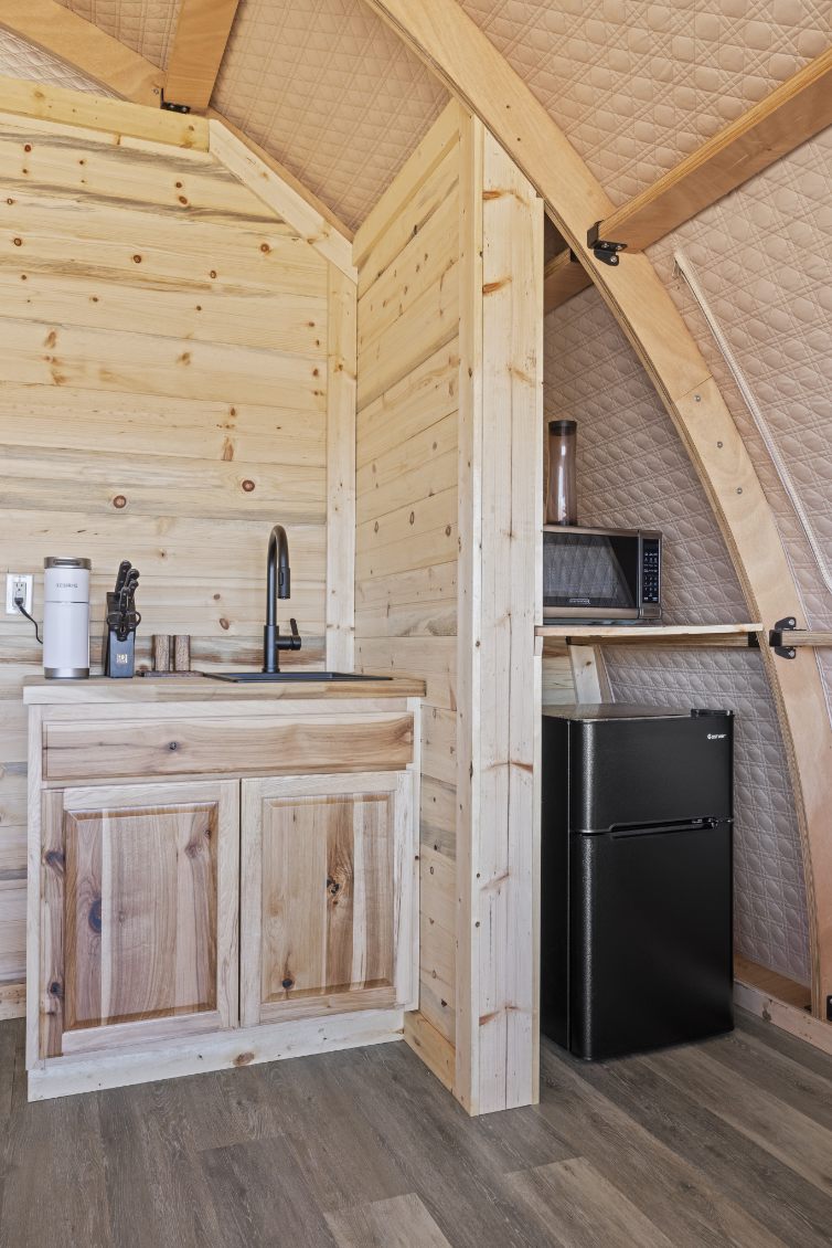 Alt text: "Interior view of a wooden glamping pod featuring a kitchenette with a sink, cabinets, and a small refrigerator. The stylish design highlights the use of natural wood, ideal for sustainable living or a luxury Airbnb experience. Perfect for those interested in geodesic dome house kits or backyard dome solutions."
