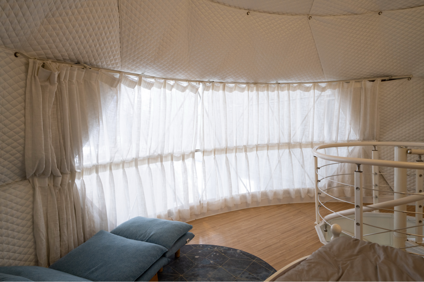 Interior view of a luxurious geodesic dome with quilted walls and large windows draped with sheer curtains, featuring a cozy seating area and a spiral staircase. Ideal for glamping, this dome offers a stylish and comfortable space for relaxation or as a unique backyard office. Keywords: geodesic dome, luxury glamping, dome home, artemis pod.