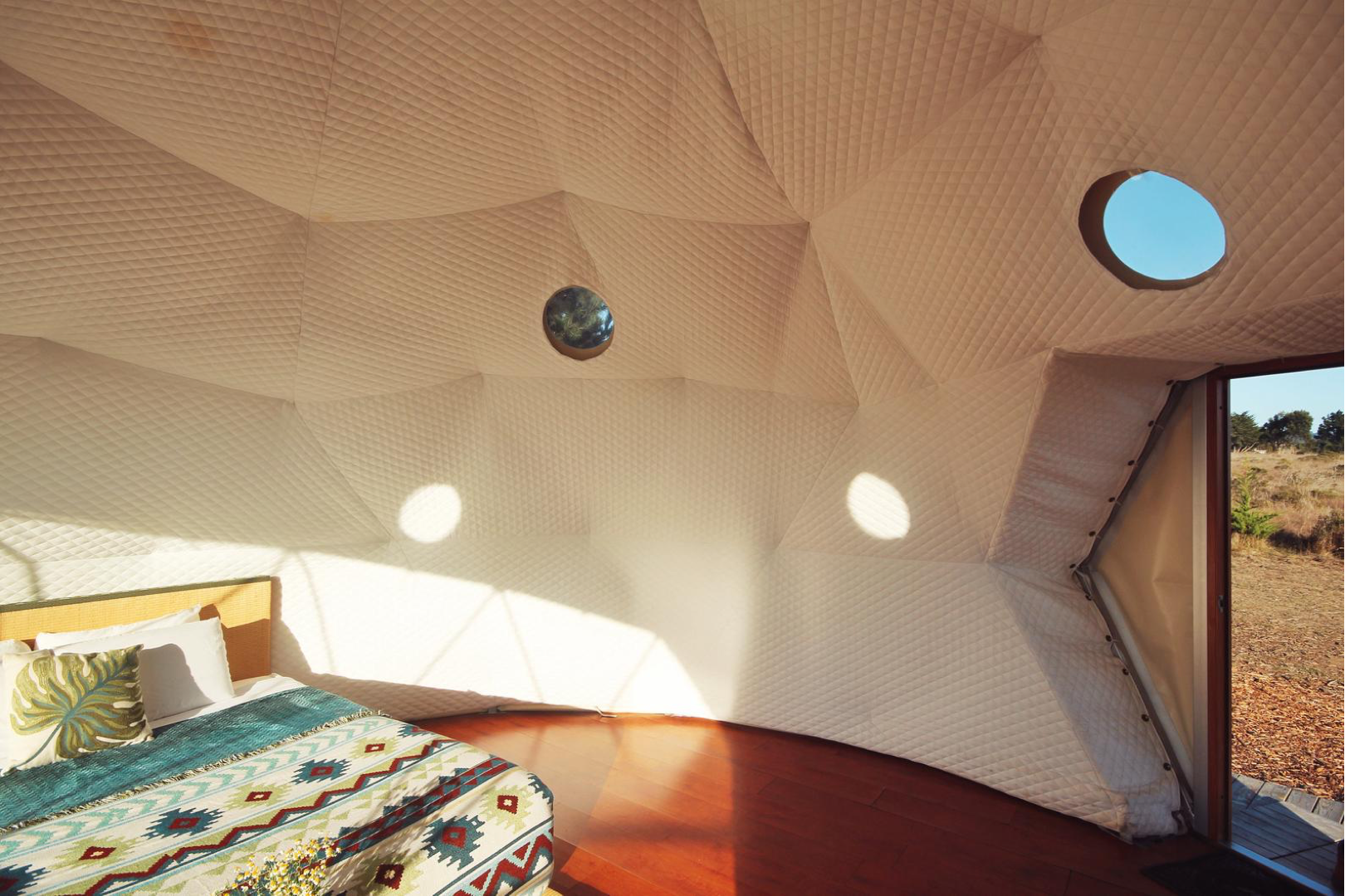 Interior view of a stylish geodesic dome with porthole windows, featuring a cozy bed with patterned bedding. Ideal for luxury glamping or as a unique backyard dome office.