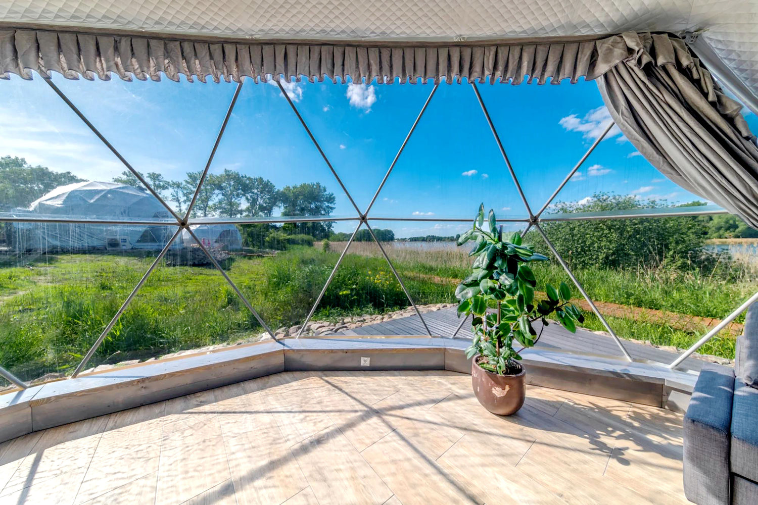 Alt text: &quot;Interior view of a geodesic dome with large porthole windows, showcasing a scenic outdoor landscape. The dome features a stylish interior with a potted plant and wooden flooring, ideal for glamping or use as a luxury Airbnb. Keywords: geodesic dome, porthole windows, glamping, luxury Airbnb, dome home.&quot;