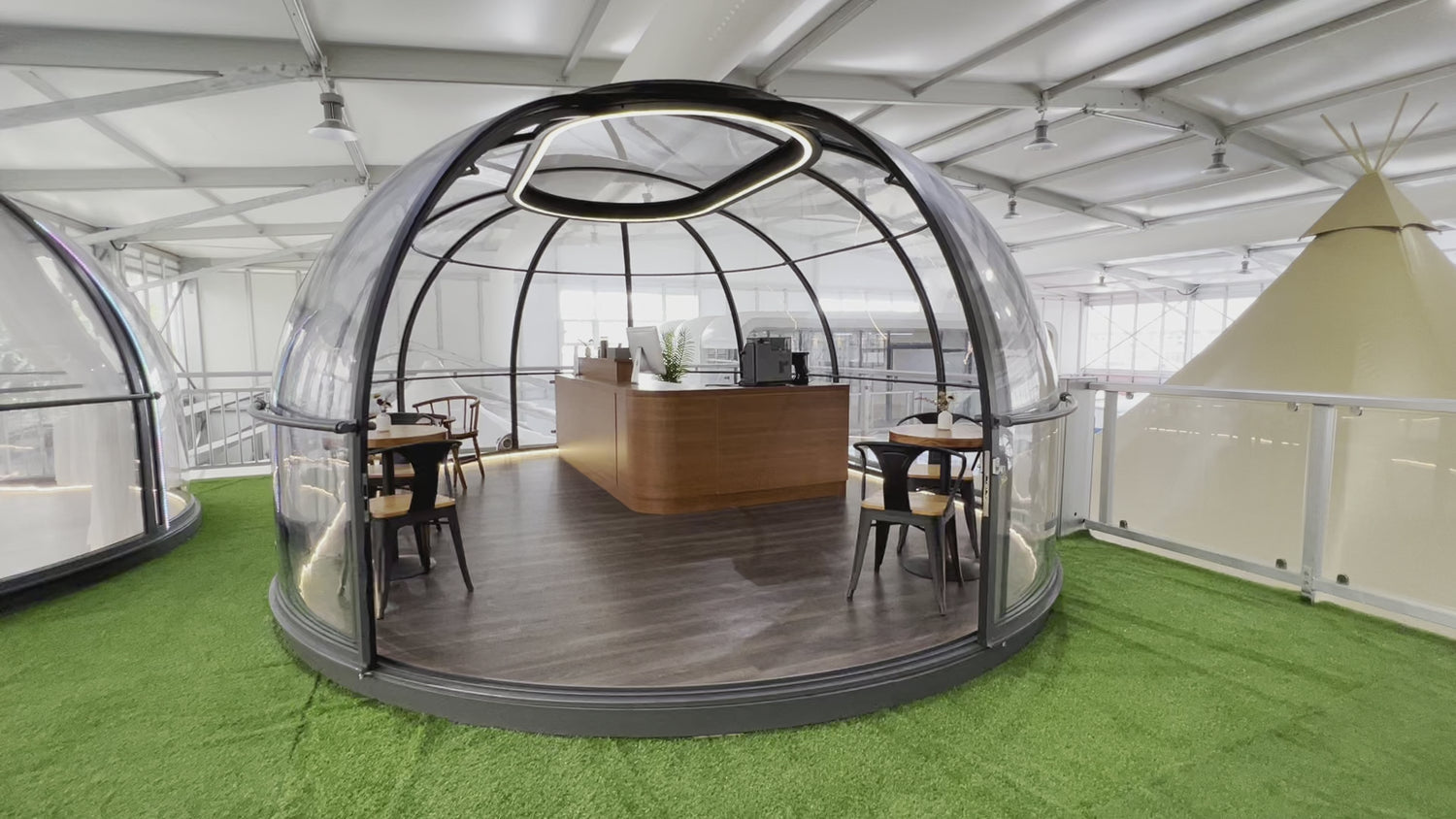 Alt text: &quot;Modern geodesic dome with transparent walls, featuring a stylish interior setup with tables and chairs, ideal for glamping or a backyard office. The dome is situated on a green artificial grass surface, showcasing the company&