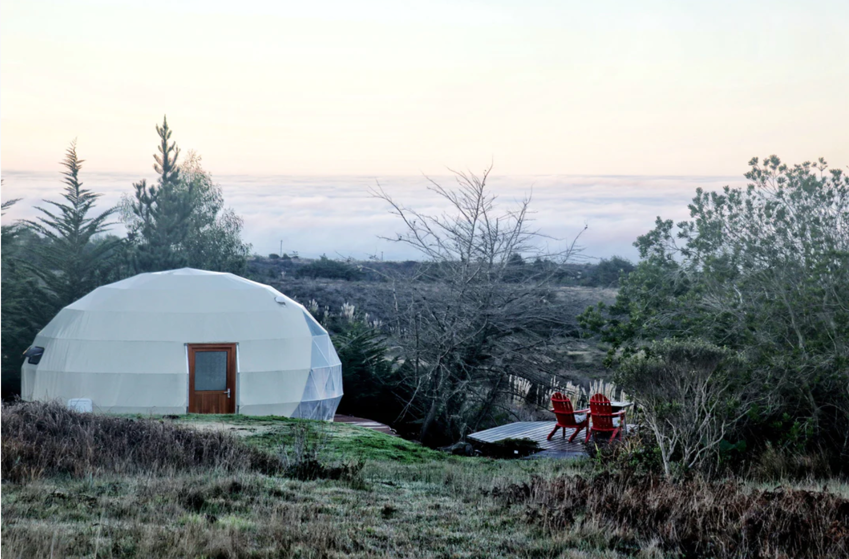 Alt text: &quot;Geodesic dome house with porthole windows set in a scenic outdoor landscape, featuring red chairs on a wooden deck. Ideal for glamping, sustainable living, or as a luxury Airbnb option. High-quality dome home solution by a company specializing in glamping pods and geodesic dome kits.&quot;